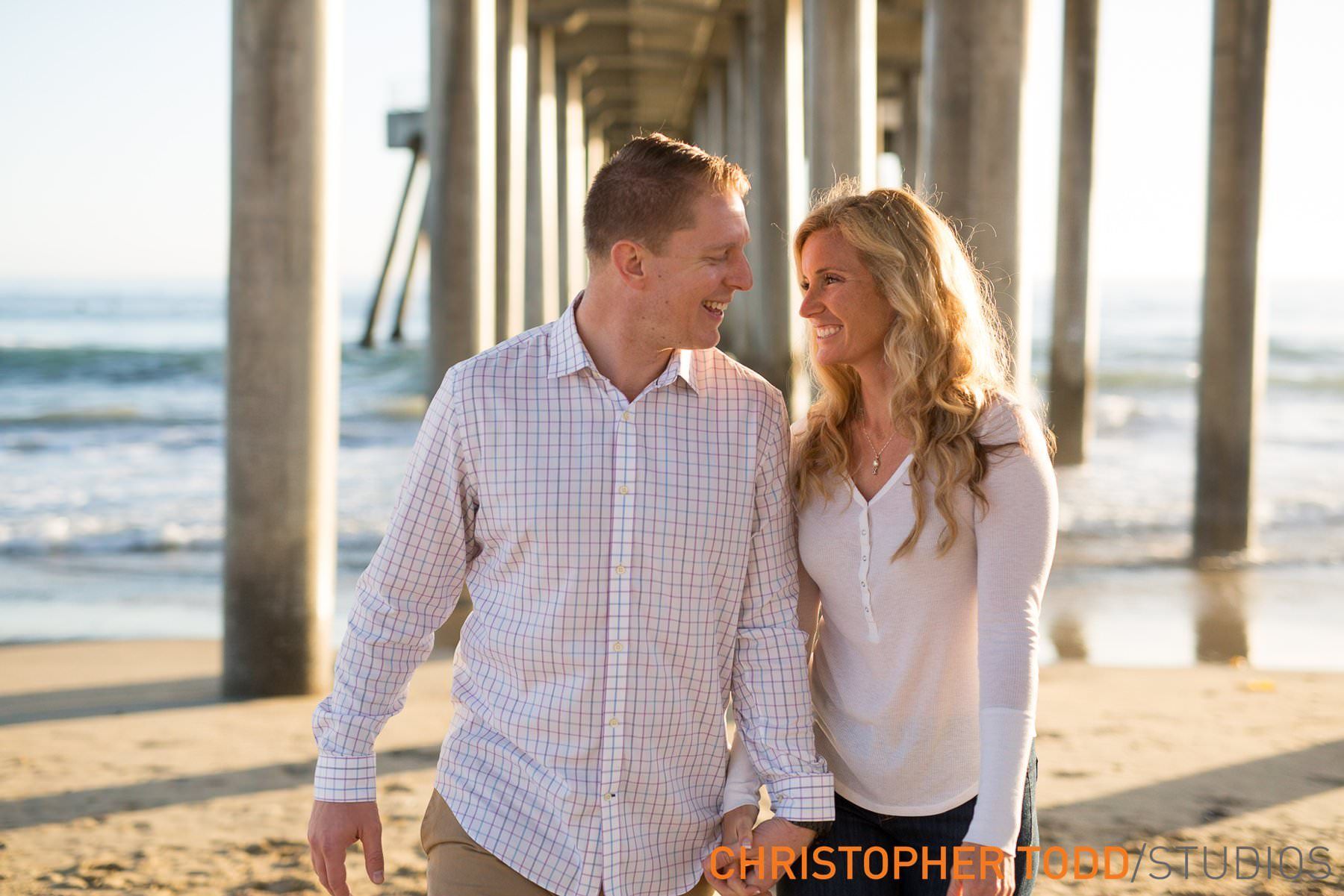 Couple holding hands, smiling at each other on a beach under a pier.
