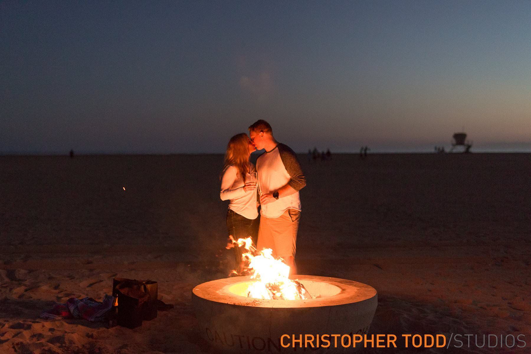 A man and woman are kissing in front of a fire pit on Huntington beach.