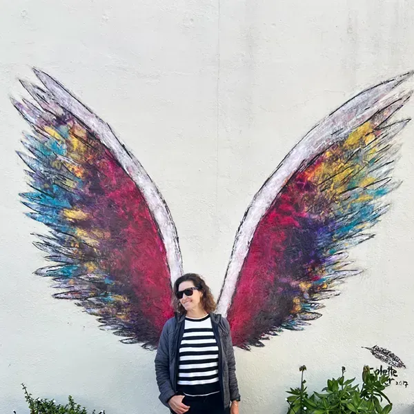 A woman is standing in front of a painting of wings on a wall painted by Colette