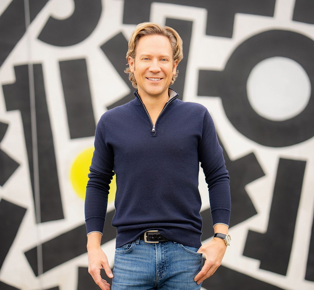 A man in a blue sweater and jeans is standing in front of a wall that has the letter o on it