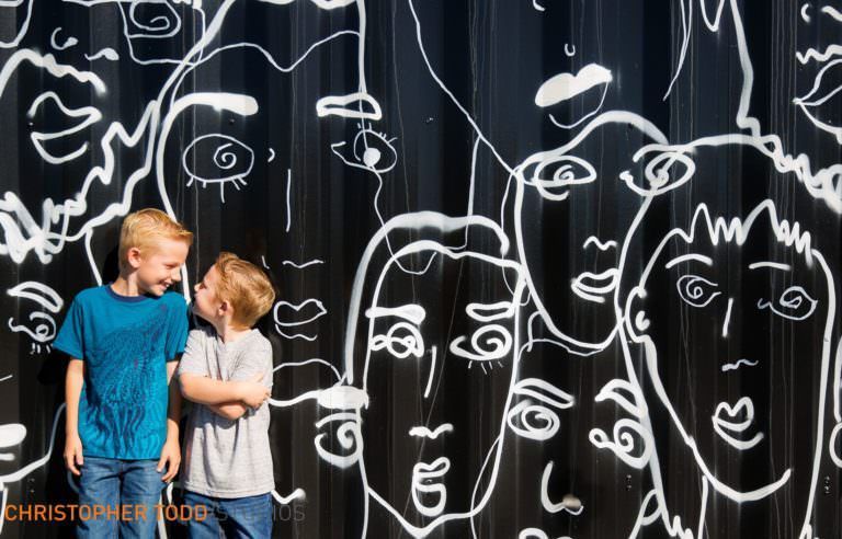 Two boys are standing in front of a wall with drawings of faces on it.