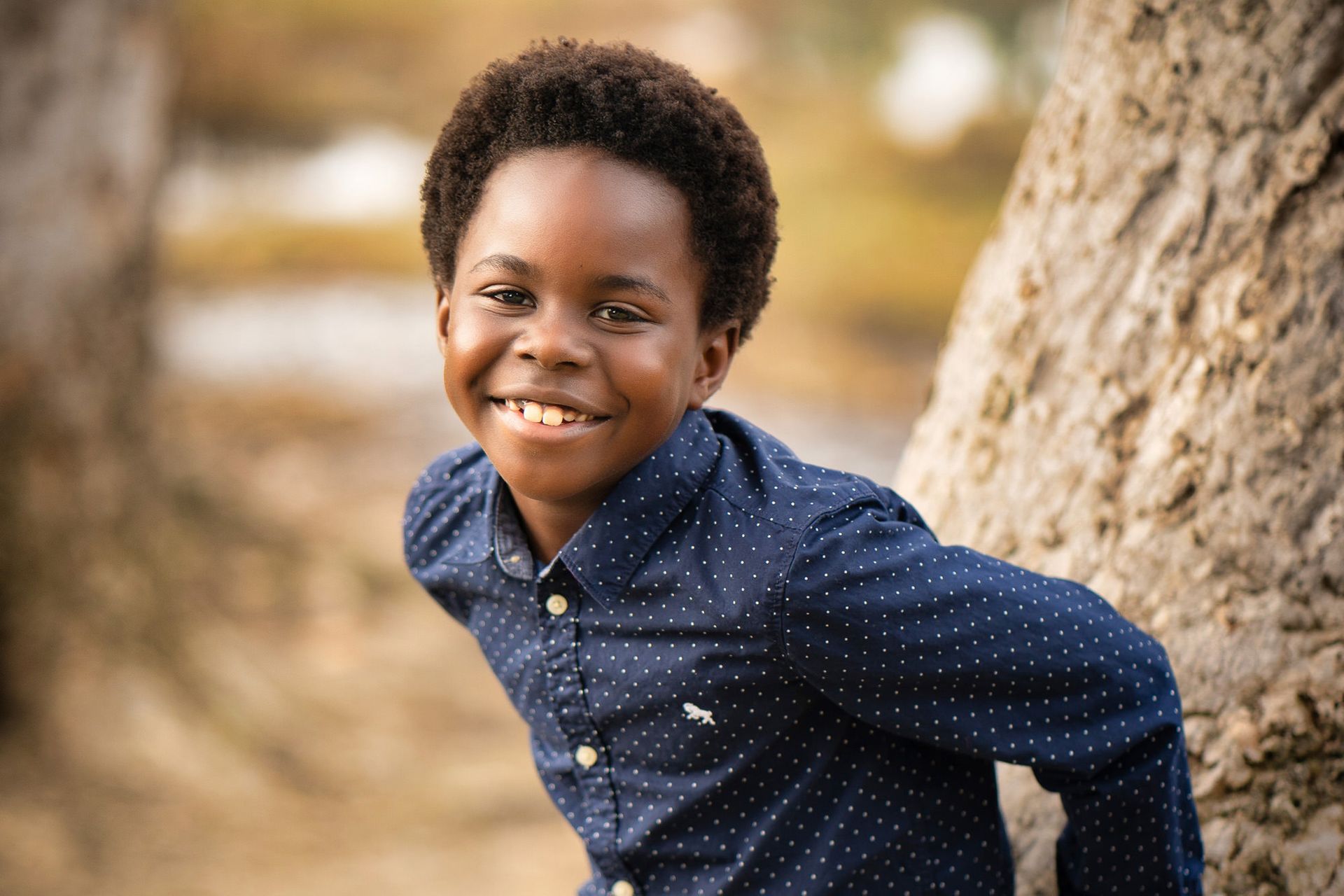 A young boy in a blue shirt is leaning against a tree and smiling in Huntington Beach Park. 