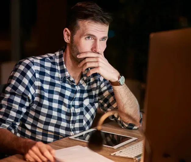 Corporate Team Headshot Studio Man in plaid shirt thoughtfully looking at computer screen, hand to chin, at a desk with tablet and lamp.