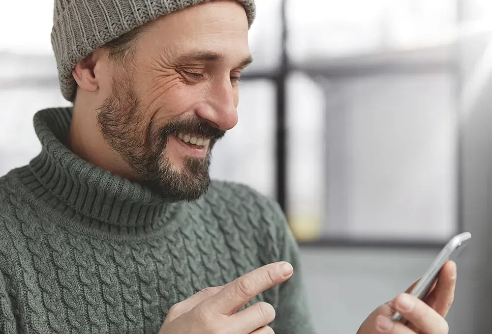 A man wearing a hat and sweater is using a cell phone.