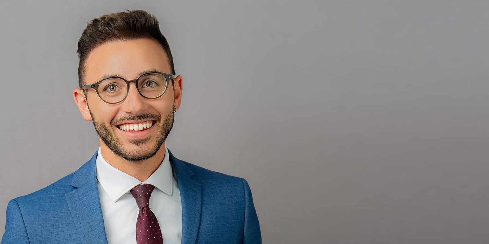 Male Corporate Team Headshots Man wearing glasses, smiling, in a blue suit and maroon tie against a gray background.