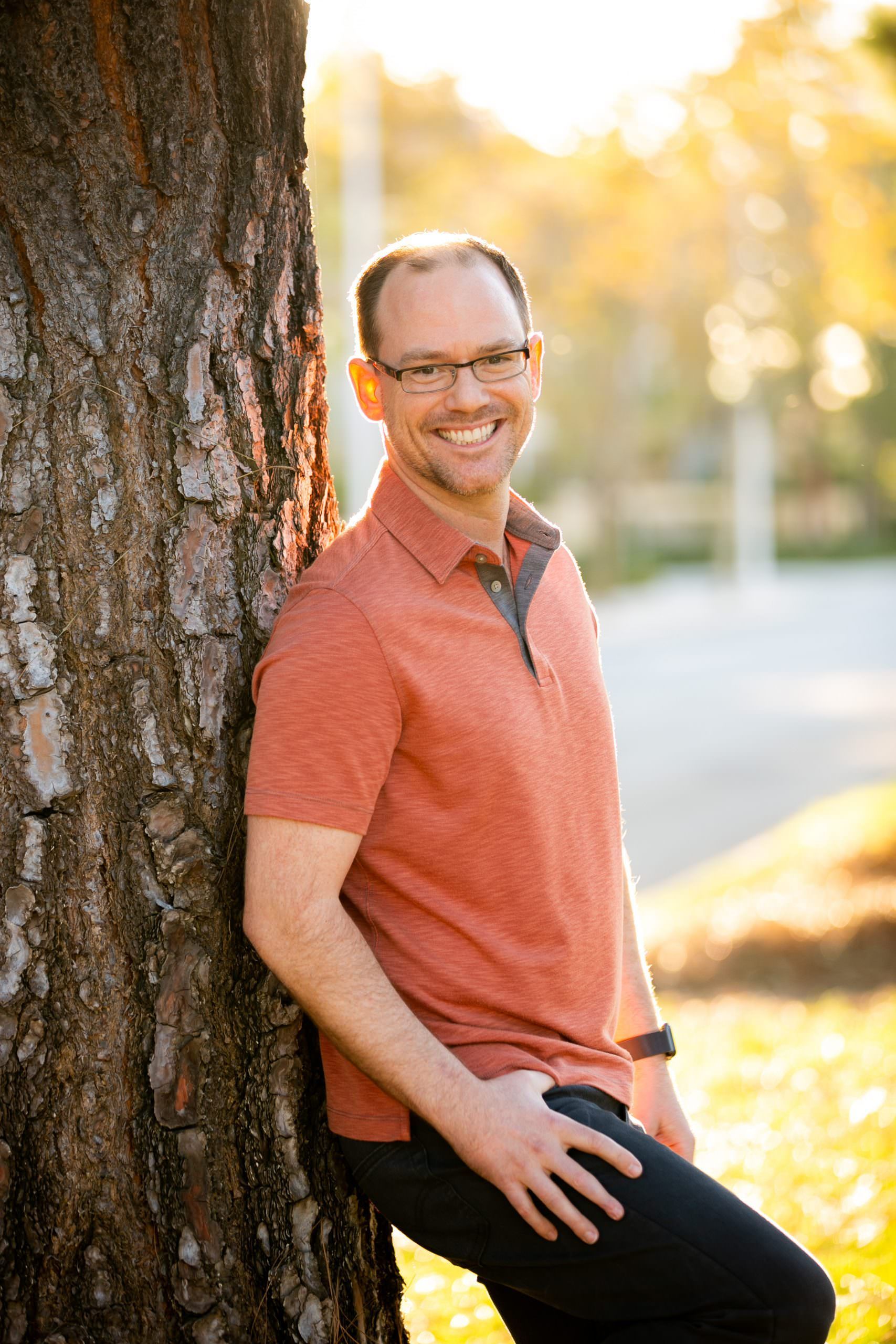 A man is leaning against a tree and smiling for a dating app