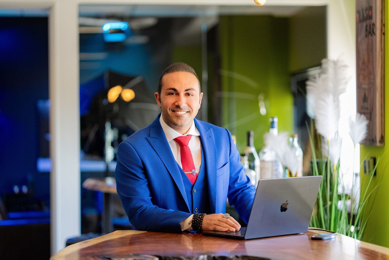 Attorney Headshots Irvine Ca. A smiling person in a blue suit and red tie sitting at a wooden table with a laptop in a brightly lit office.