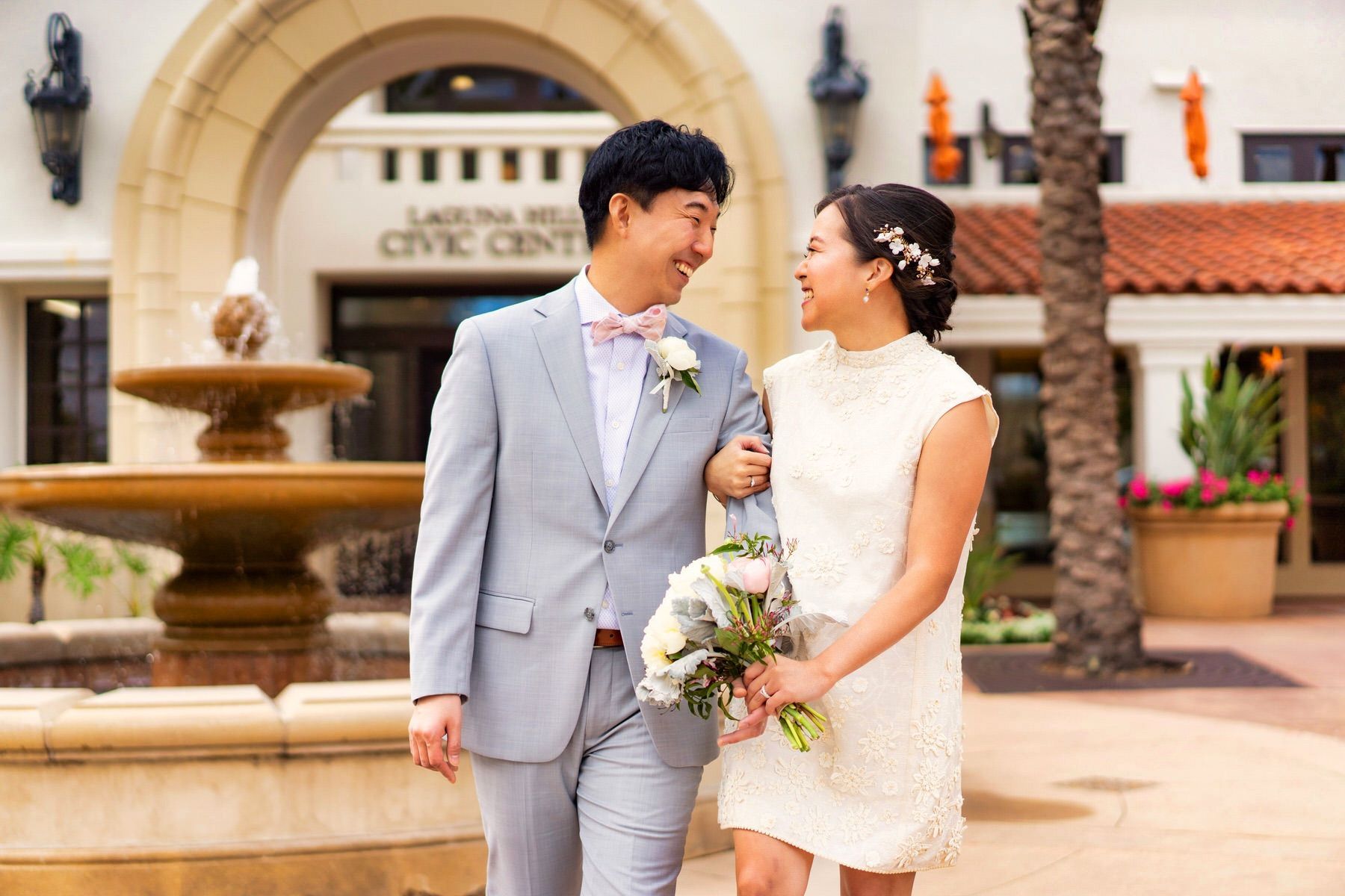 A bride and groom are walking in front of a fountain.