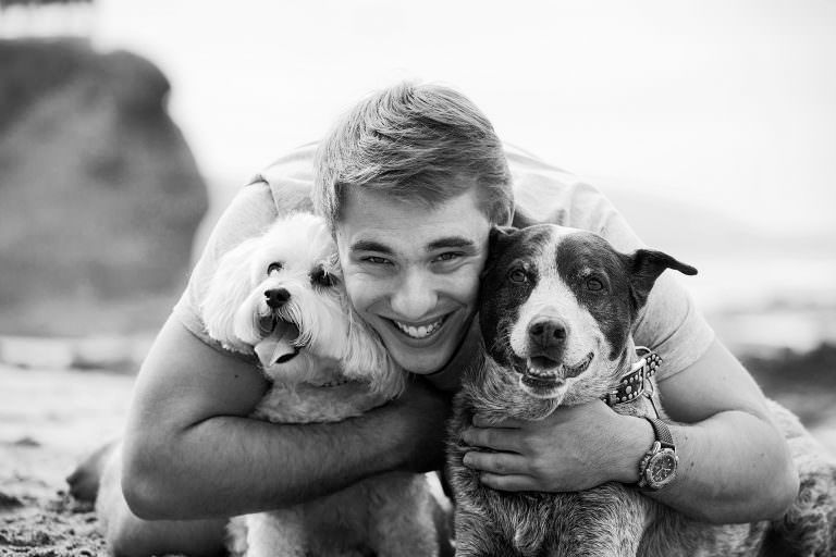 Ayoung man is hugging two dogs on the beach in a black and white photo.