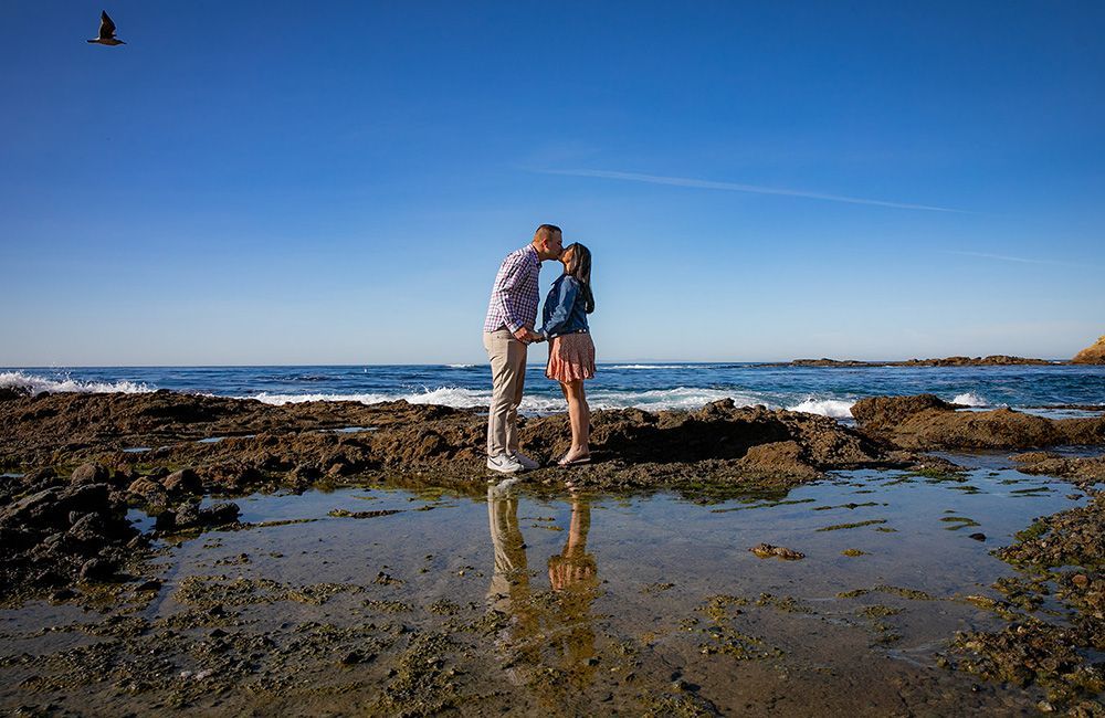 Surprise Marriage Proposal Photographer A man and a woman are kissing on a rocky beach near the ocean.
