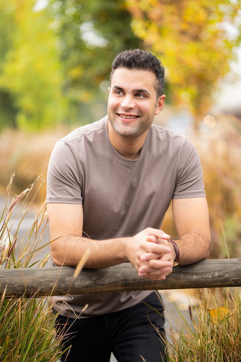 A man is leaning on a wooden fence and smiling for a professional dating photo.