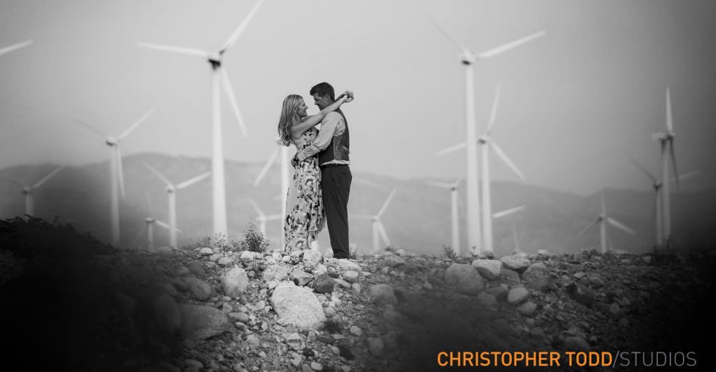 A black and white photo of a man and woman hugging in front of windmills in Palm springs