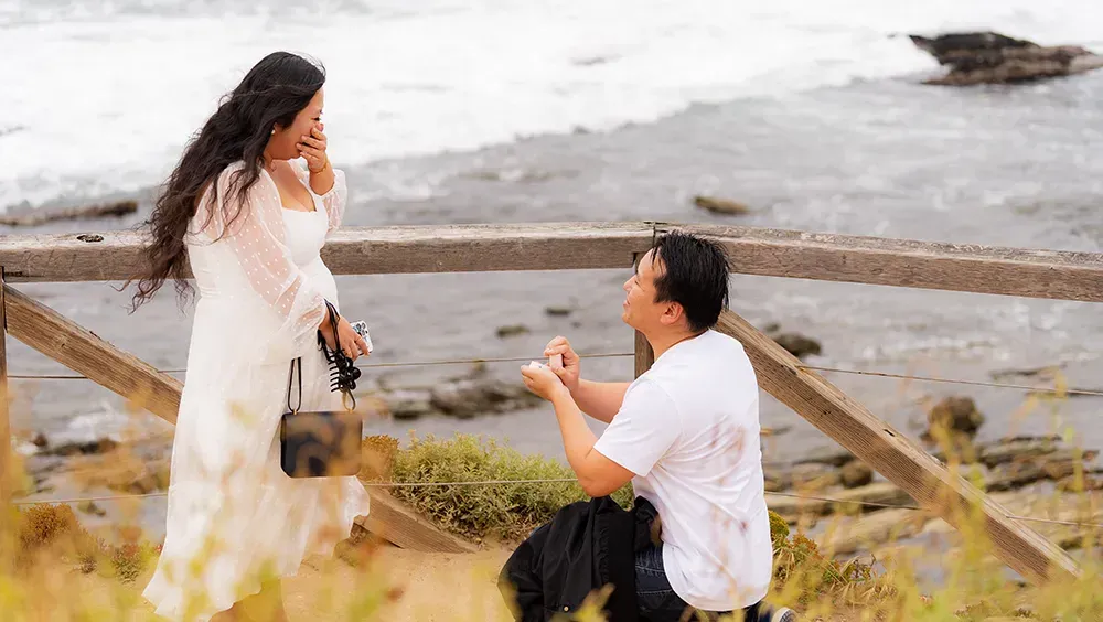 A man is kneeling down and proposing to a woman on the beach at Crystal Cove State Park.