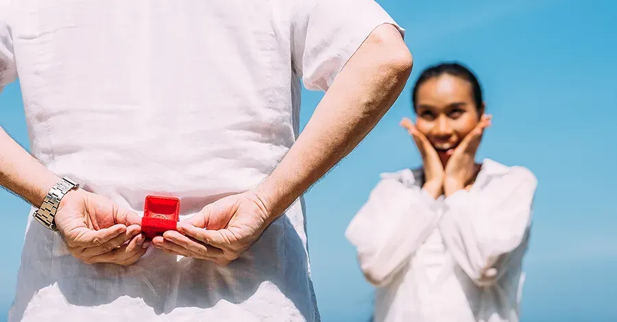 A man is holding an engagement ring in a red box behind his back.