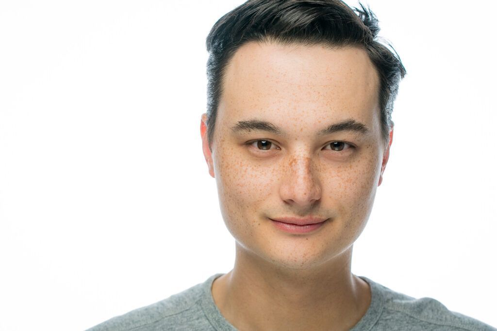 A young male actor in a grey shirt is smiling for the camera.