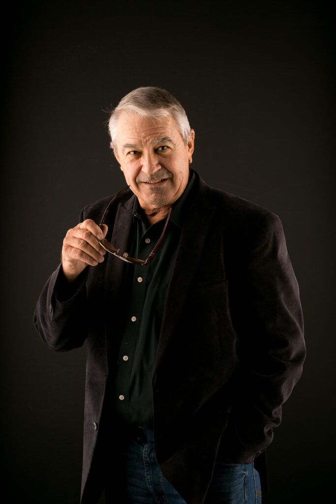 An older Actor in a black jacket is holding a pair of glasses over his ear.