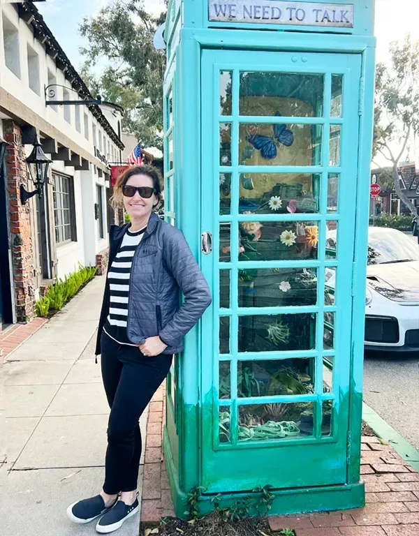 A woman is standing next to a phone booth that says `` we need to talk ''. Near main beach in Laguna.
