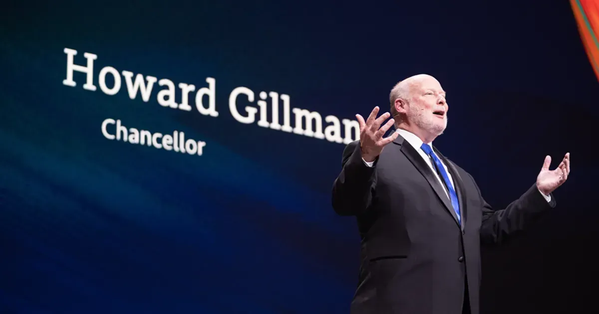 Howard Gillman speaking on stage beside projected name text, arms raised, in a dark blue-lit setting