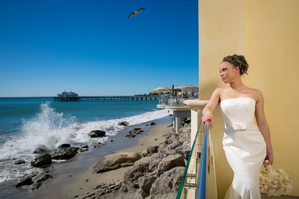 A Malibu bride in a white dress is standing on a balcony overlooking the ocean.