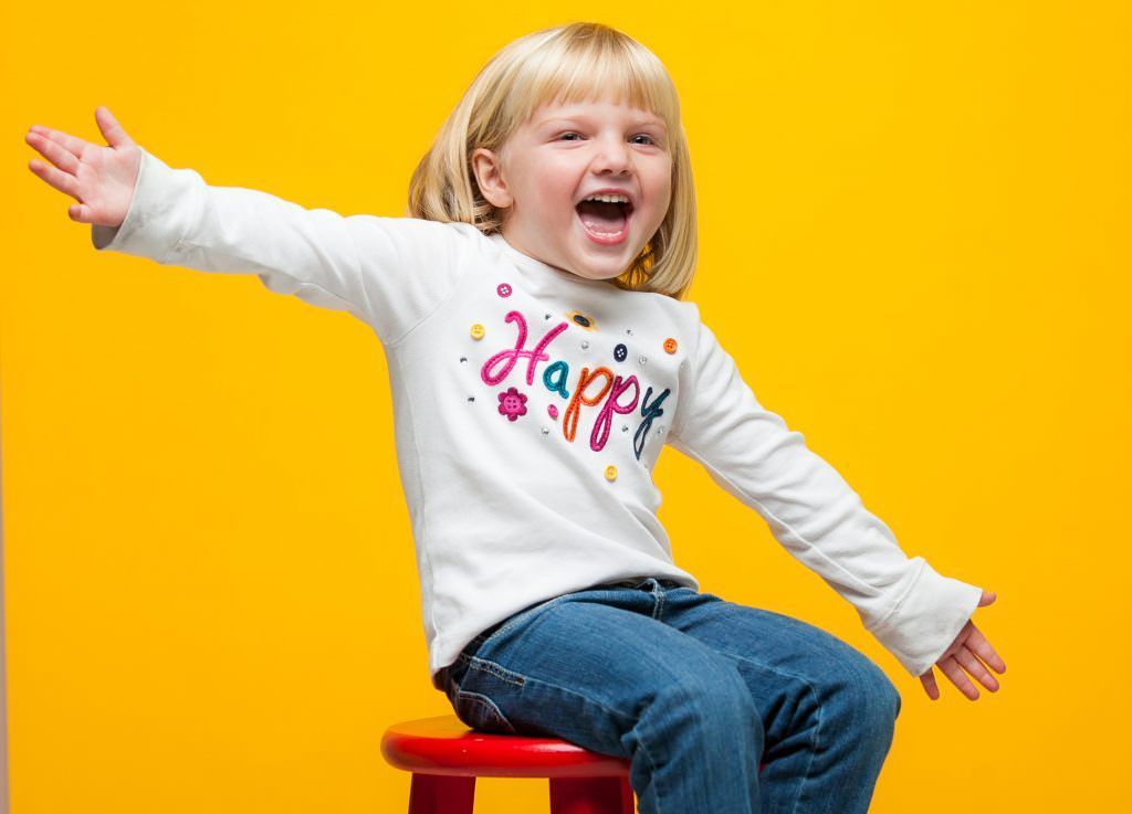 Fun Studio Portraits A little girl is sitting on a red stool with her arms outstretched.