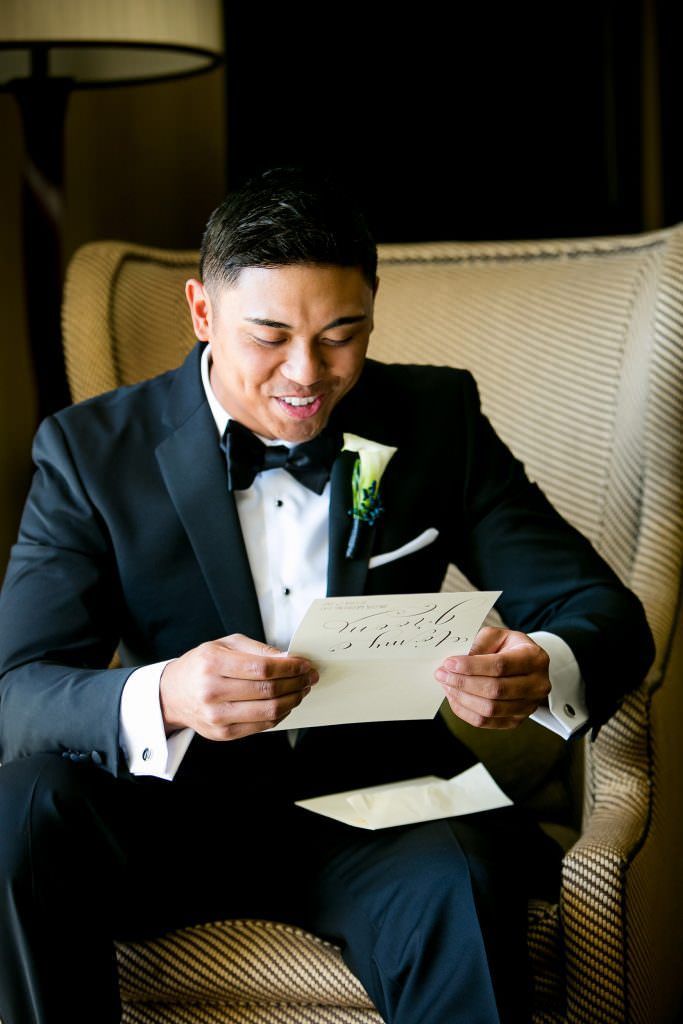 A groom in a tuxedo is sitting in a chair reading a letter from his bride.