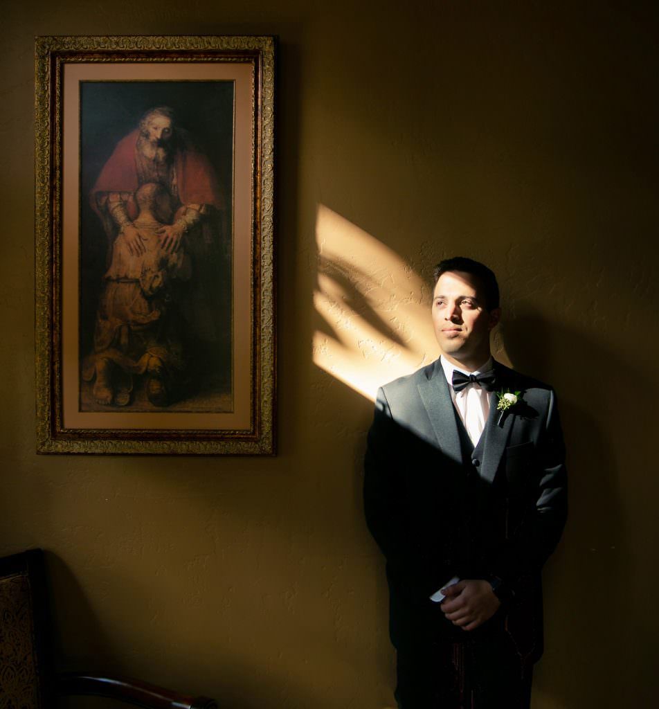 A Catholic groom in a suit and bow tie is standing in front of a painting on a wall.