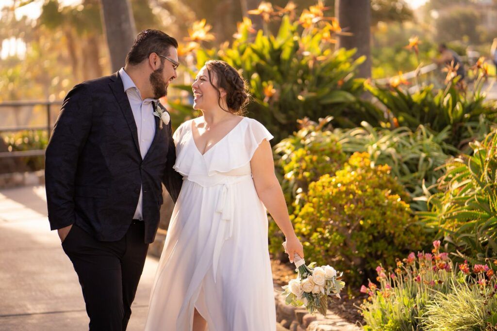 Heisler park Laguna Beach elopement A bride and groom are walking down a sidewalk in a garden in Heisler Park Laguna Beach.