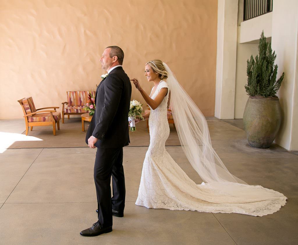 A bride wearing a long veil taps her groom on the shoulder during a first look in Huntington Beach