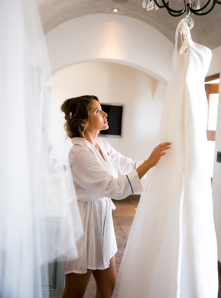 A woman in a white robe is looking at her wedding dress hanging on a chandelier.