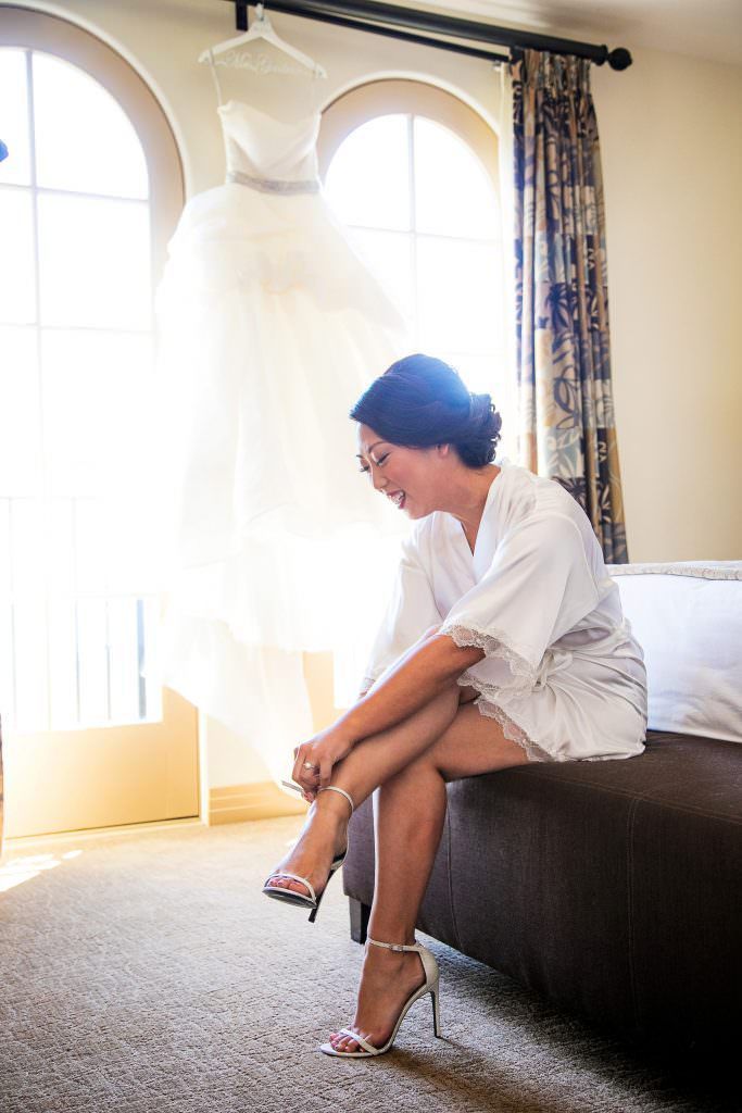 A bride is sitting on a bed putting on her wedding shoes.