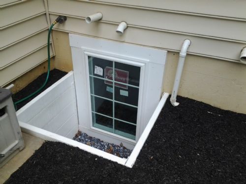 A basement window with a white frame set in a gravel-filled egress well against a light-colored, siding-covered house.