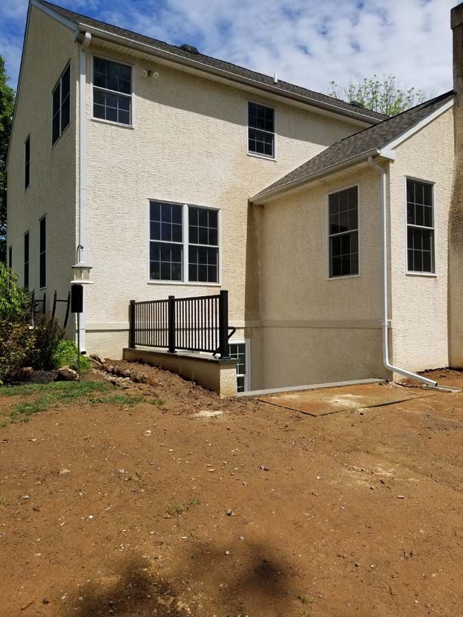 A beige, two-story house with a basement window well, black railing, and freshly disturbed soil in the yard.