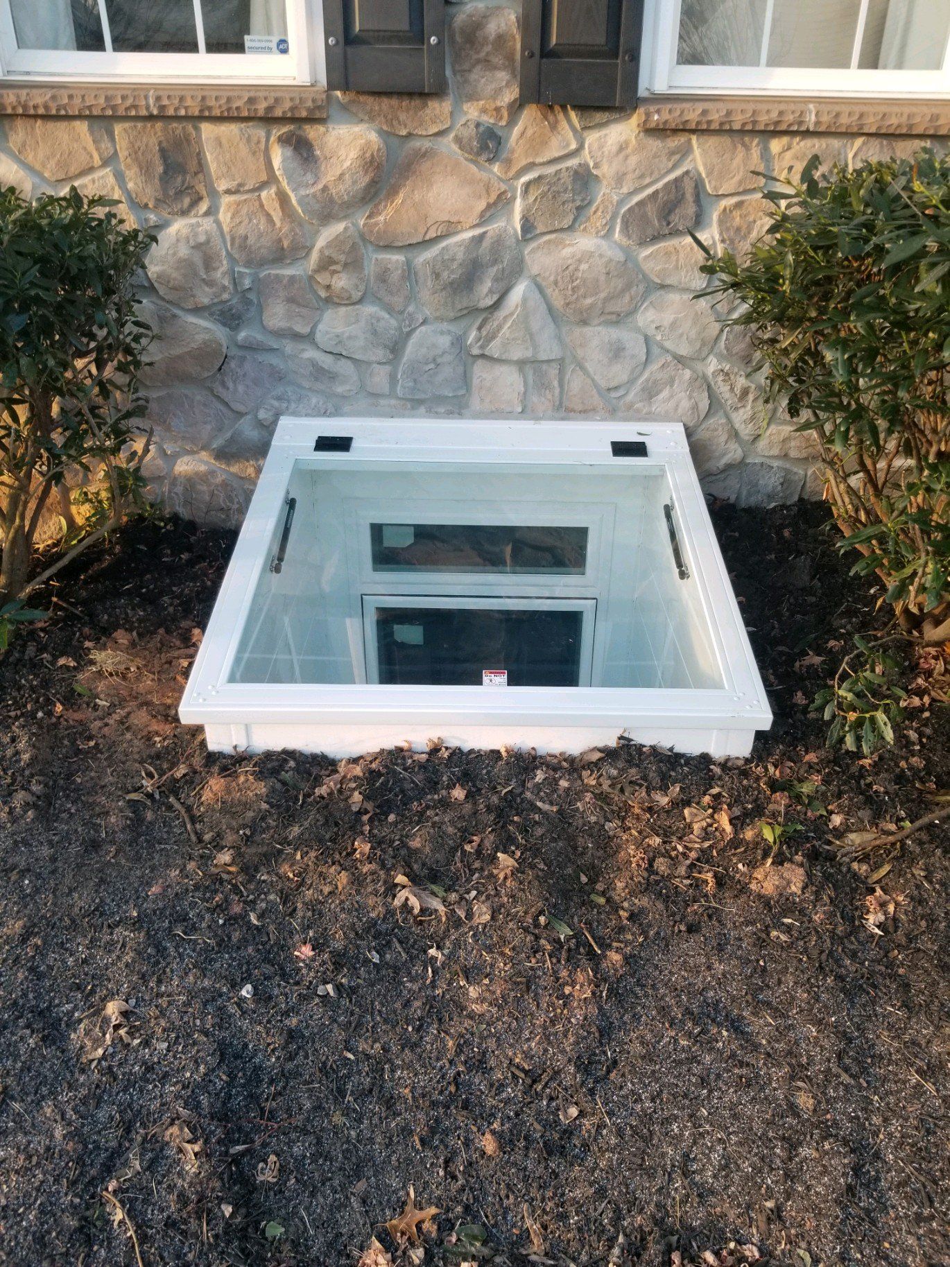 A clear, rectangular basement window well cover sits on dark mulch against a stone house exterior.