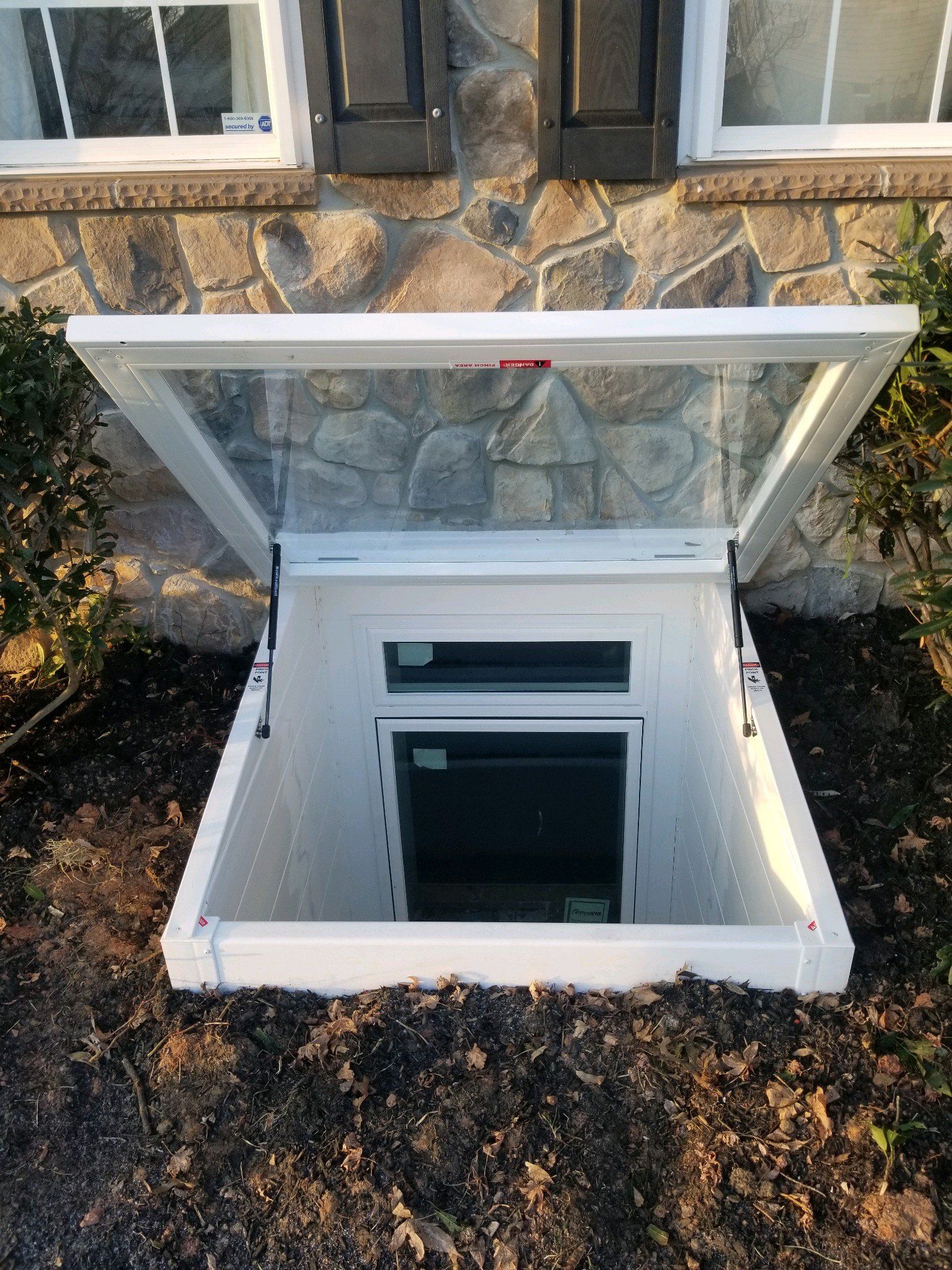 A white window well cover is propped open, revealing an egress window set into a stone basement wall surrounded by mulch.