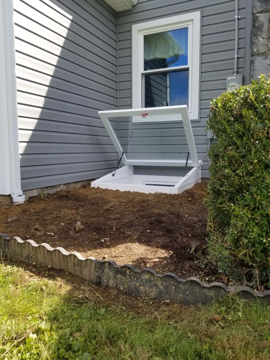 A white basement egress window well cover stands open against the side of a gray-sided house in a dirt garden bed.