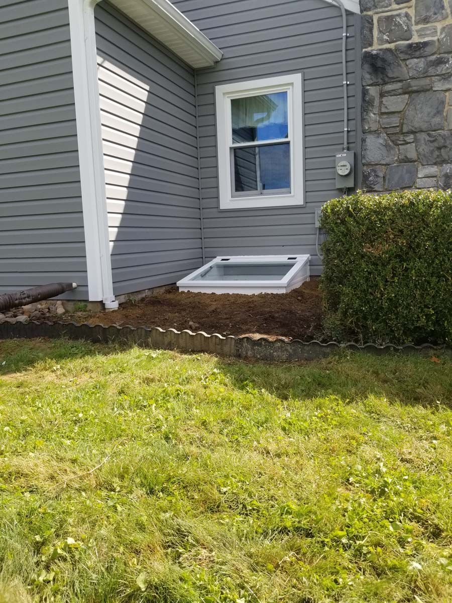A gray-sided house with a white-framed window above a basement window well, set near a stone wall and a green hedge.