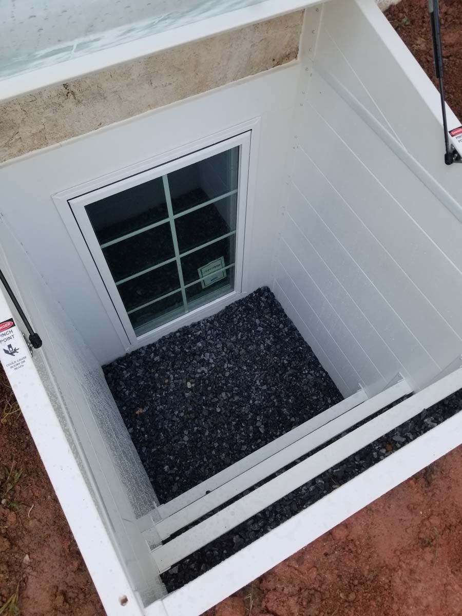A white basement window well with a grid-patterned window, gravel base, and an open, hinged transparent cover.