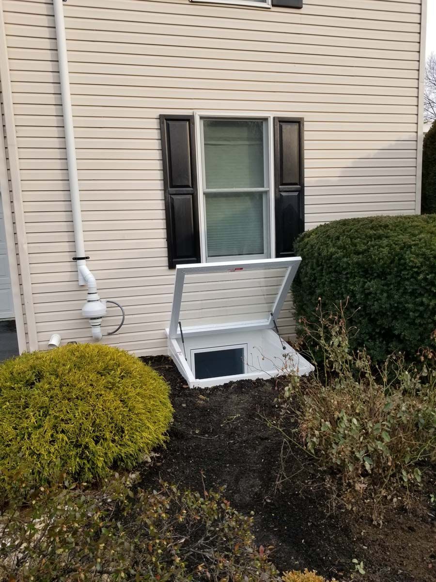 A white window well cover is propped open against the beige vinyl siding of a house, beneath a window with black shutters.