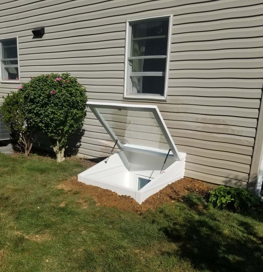 A white basement window well cover with a clear panel is open against the tan siding of a house.