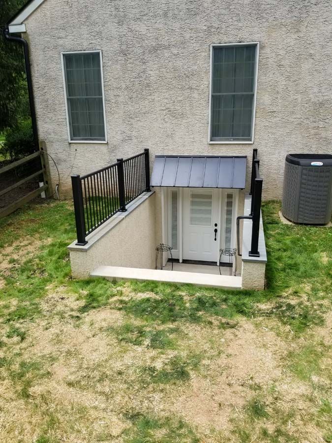 A basement walkout entrance with a white door, metal awning, and black railings set into the exterior of a stucco home.