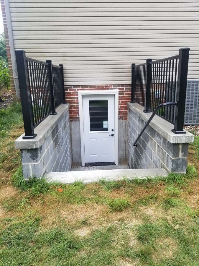 An outdoor basement entry with a white door, concrete walls, and black metal railings leading down from the yard.