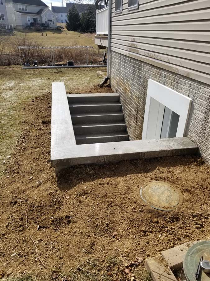 A concrete basement stairwell entrance leading down to a white-framed door on the side of a house with tan siding.