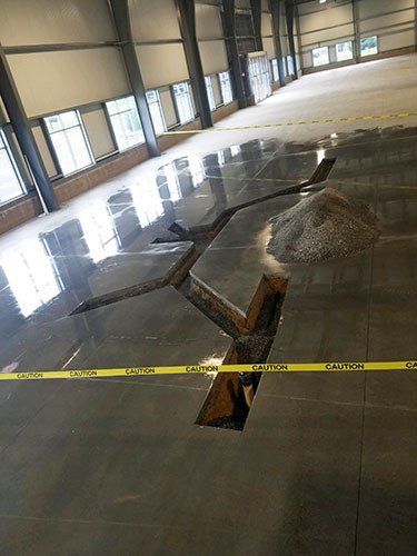 Concrete floor inside a large, empty warehouse with trenches cut into the surface and yellow caution tape across them.