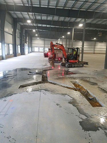 An orange excavator works inside a large industrial warehouse, where concrete floor sections have been cut out.