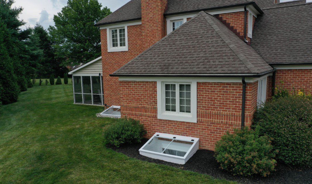 A red brick house exterior features a white-framed basement window well cover on the lawn near landscaped bushes.