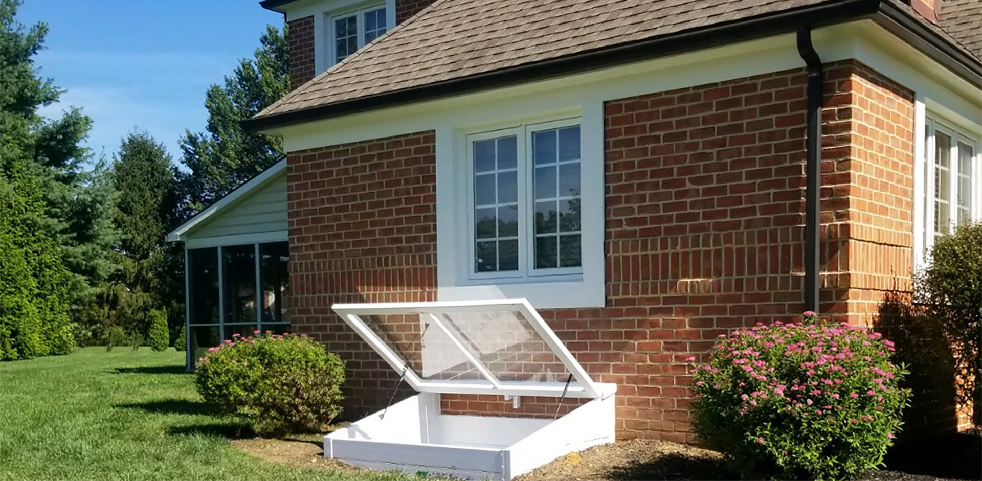 A white basement bulkhead door stands open against the brick exterior of a house surrounded by green landscaping.