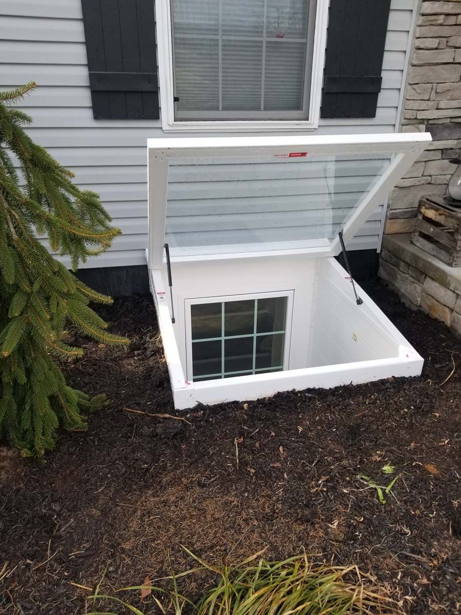 A white, hinged basement window well cover is propped open, revealing a basement window against a house exterior.