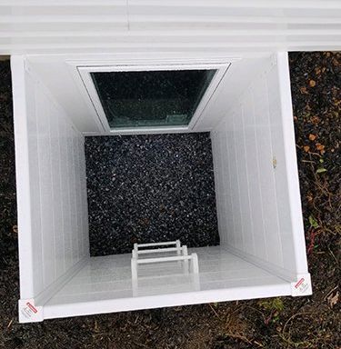 A white window well with a built-in ladder, set into the ground outside a basement window over dark gravel.