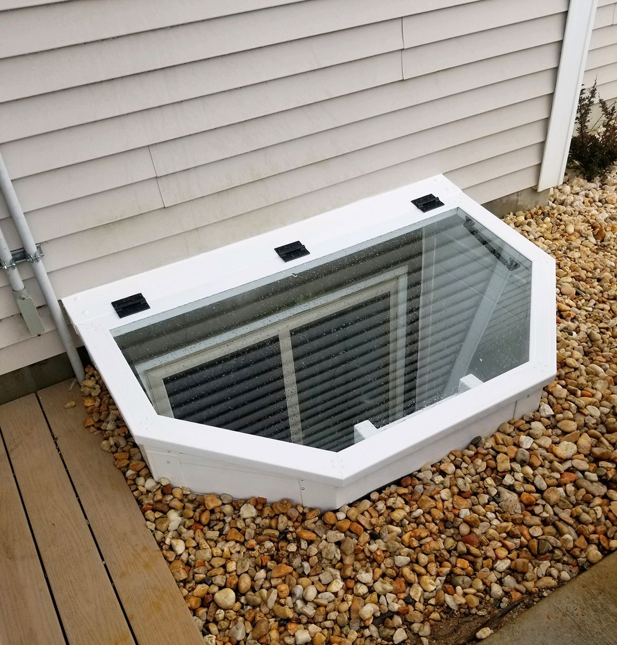A white, angled egress window cover sits over a basement window well surrounded by light-colored stones against a wall.