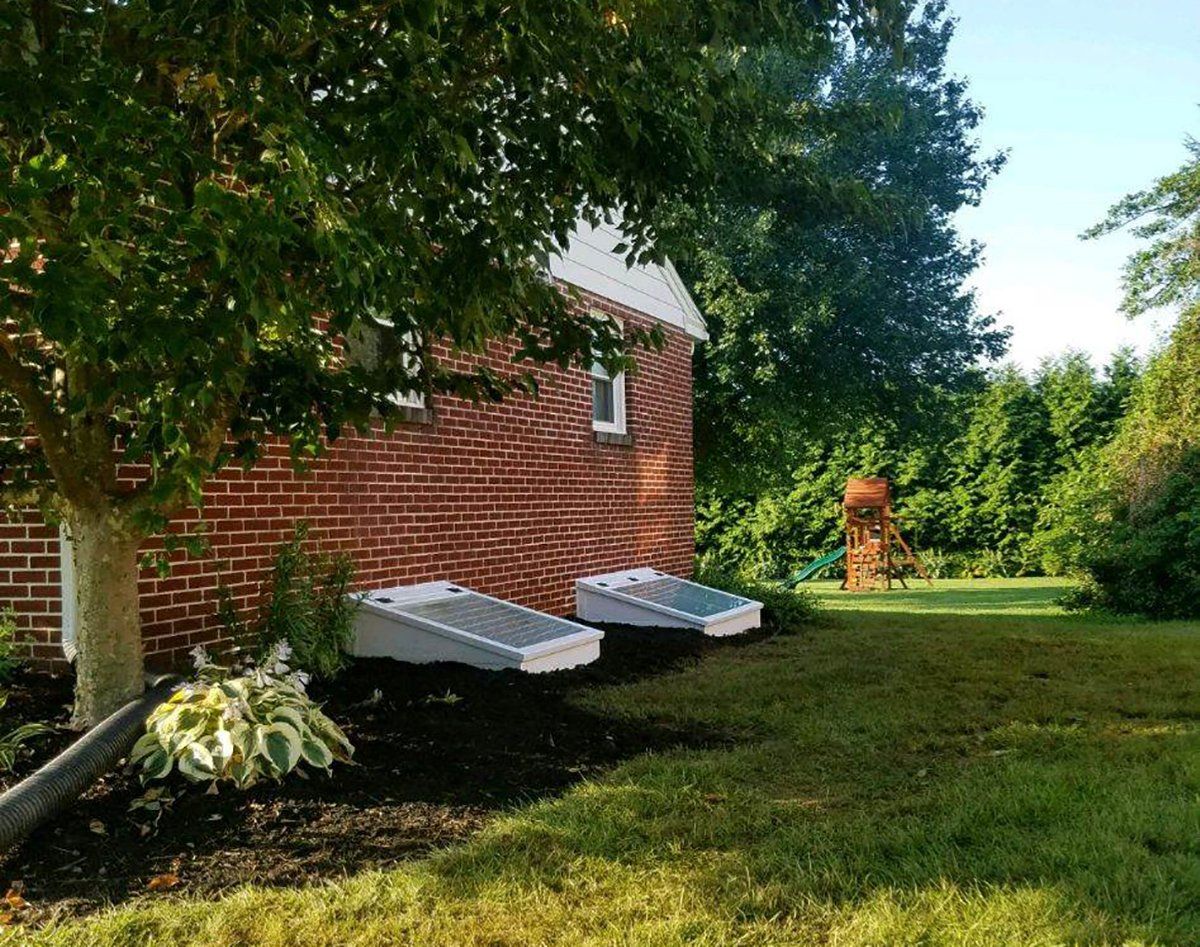 Two sloped window well covers sit against the side of a red brick house next to a tree, a garden bed, and a lawn.